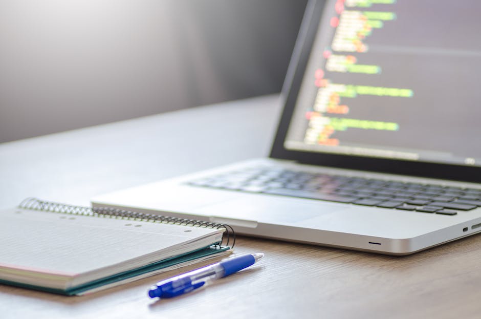 A close-up view of a clean, organized office workspace featuring an open silver laptop displaying lines of colorful code on a dark background, positioned on a wooden desk. Beside the laptop, there is 