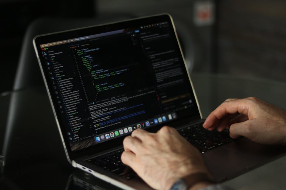A close-up view of a clean, organized office workspace featuring an open silver laptop displaying lines of colorful code on a dark background, positioned on a wooden desk. Beside the laptop, there is 